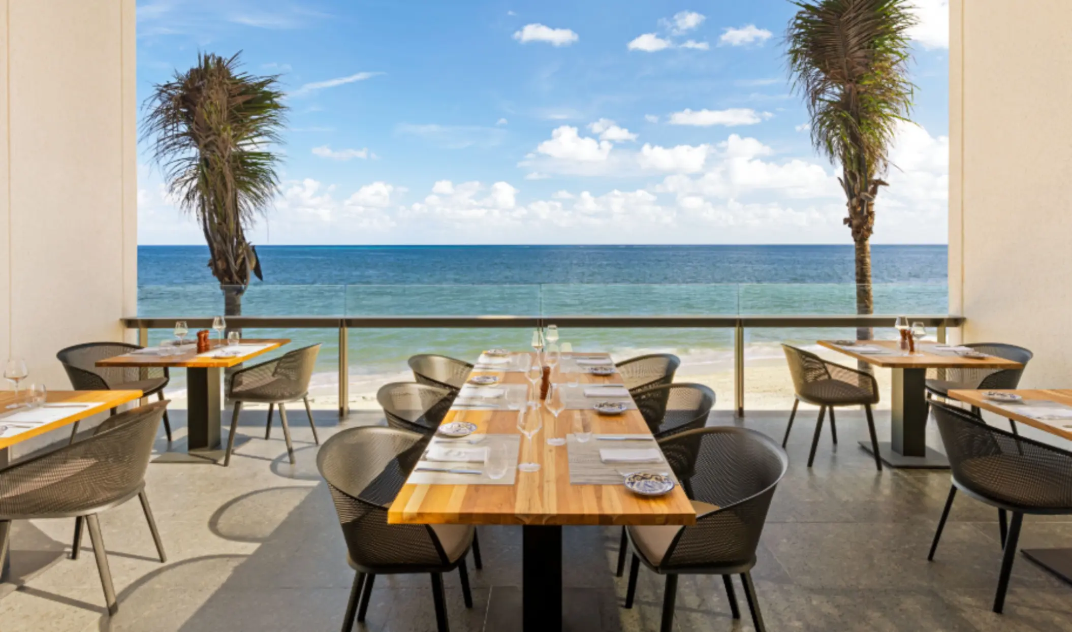 Open-air beachfront dining with wood tables, mesh chairs, and panoramic views of the ocean and palm trees.