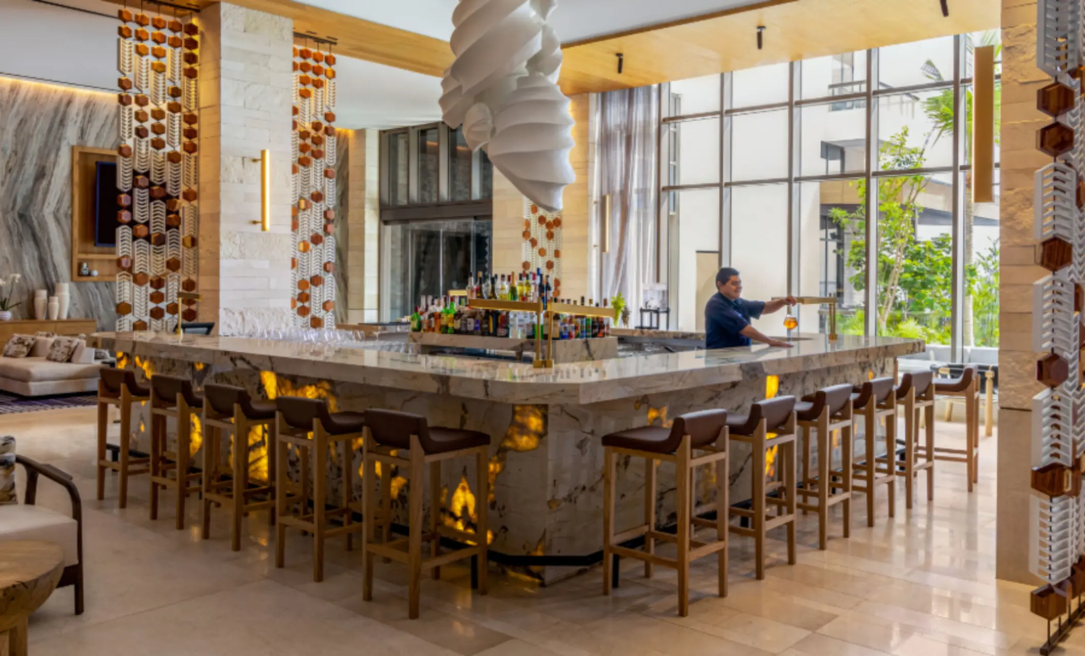 Upscale lobby bar with backlit marble counter, high stools, large windows, and a bartender preparing drinks.