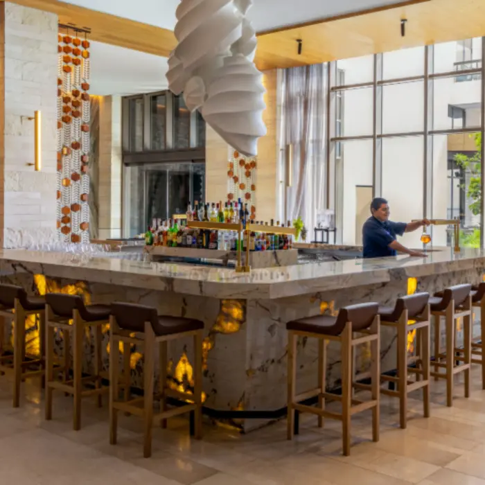 Upscale lobby bar with backlit marble counter, high stools, large windows, and a bartender preparing drinks.