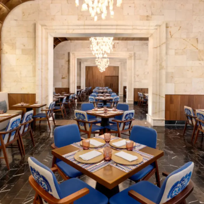 Elegant indoor dining area with blue-accent chairs, wood tables, marble floors, and chandeliers in a refined restaurant space.