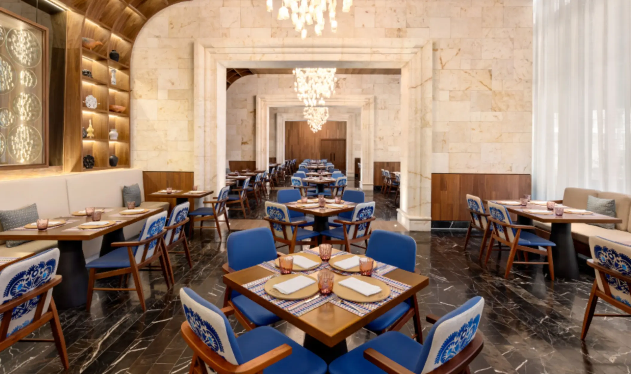 Elegant indoor dining area with blue-accent chairs, wood tables, marble floors, and chandeliers in a refined restaurant space.