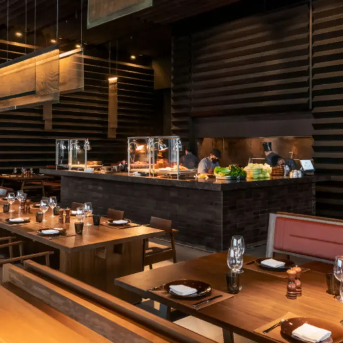 Upscale restaurant with open kitchen, wood paneling, warm lighting, and neatly arranged tables with modern place settings.