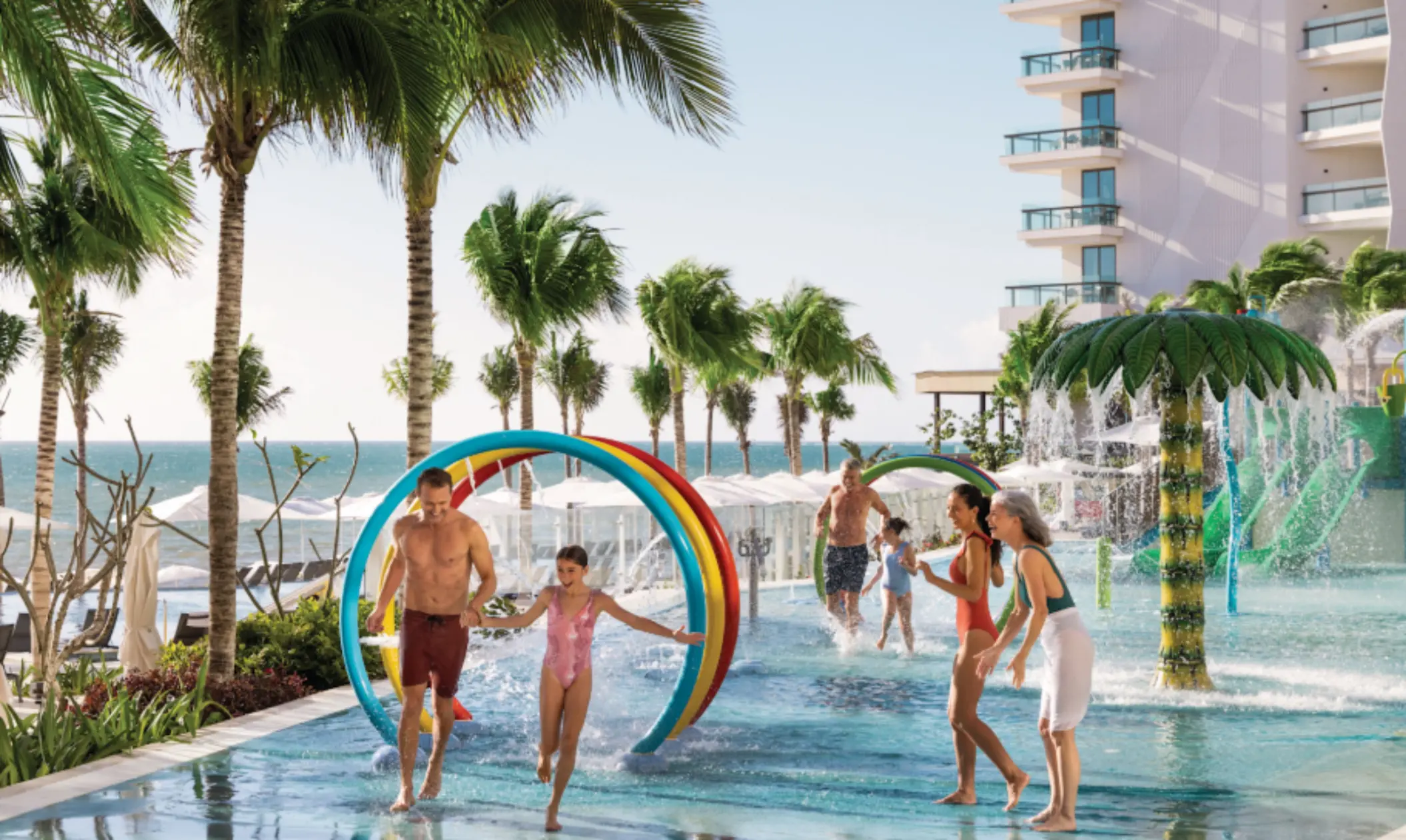 Multigenerational family playing in colorful splash pad with water arches and palm tree sprayers by the beach.