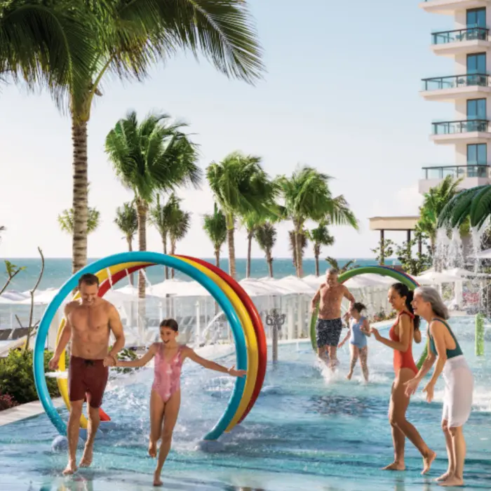 Multigenerational family playing in colorful splash pad with water arches and palm tree sprayers by the beach.