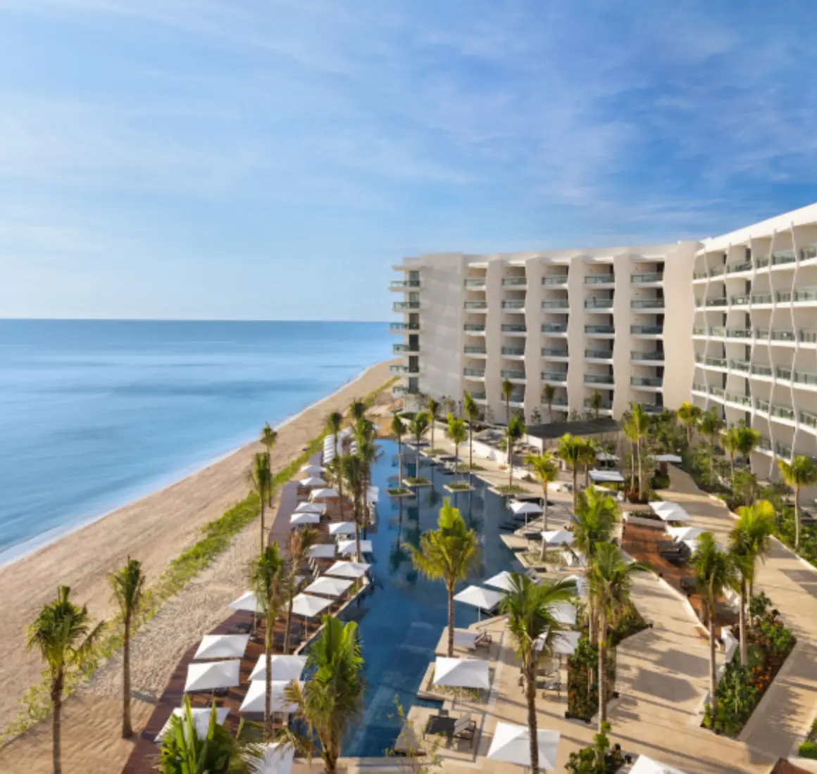 Aerial view of beachfront resort with palm trees, ocean-facing balconies, and central pool framed by loungers.