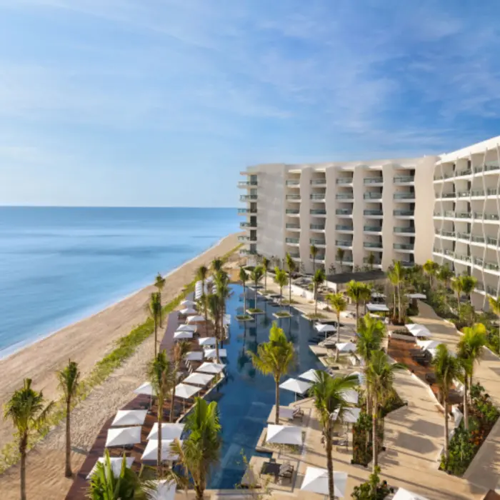 Aerial view of beachfront resort with palm trees, ocean-facing balconies, and central pool framed by loungers.