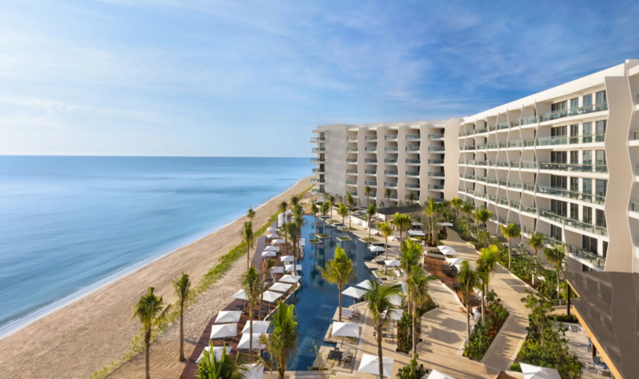 Aerial view of beachfront resort with palm trees, ocean-facing balconies, and central pool framed by loungers.