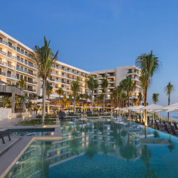 Infinity pool by the beach with resort buildings, palm trees, and sun loungers under umbrellas at sunset.