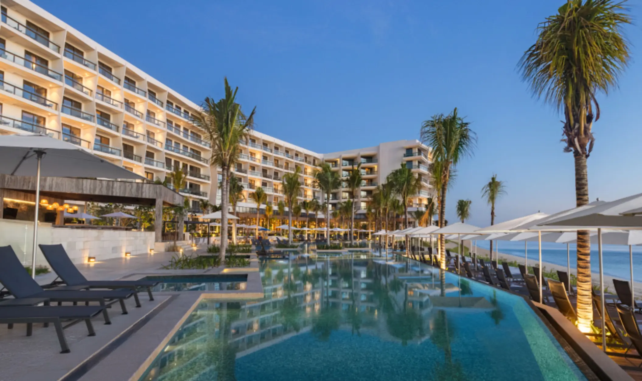 Infinity pool by the beach with resort buildings, palm trees, and sun loungers under umbrellas at sunset.