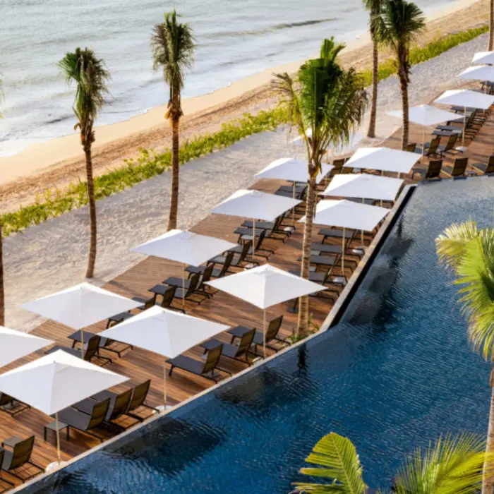 Elevated view of poolside lounge deck with rows of white umbrellas beside a calm beach and palm-lined boardwalk.