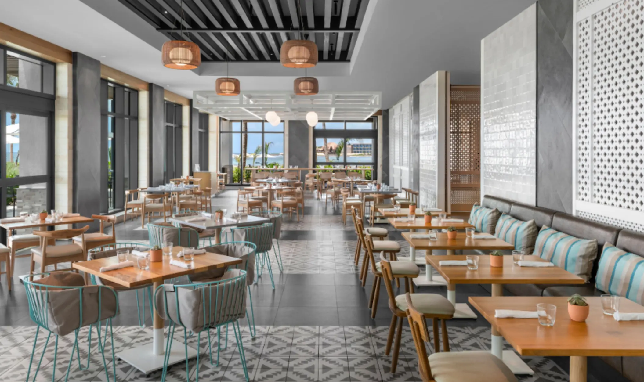Bright, coastal-style restaurant with wood tables, striped pillows, and ocean views through large windows.