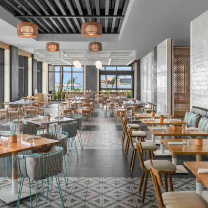 Bright, coastal-style restaurant with wood tables, striped pillows, and ocean views through large windows.
