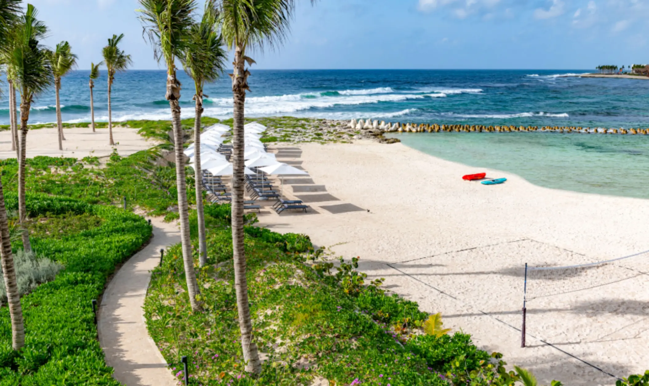 White sand beach with sun loungers and umbrellas, framed by palm trees and clear turquoise water.