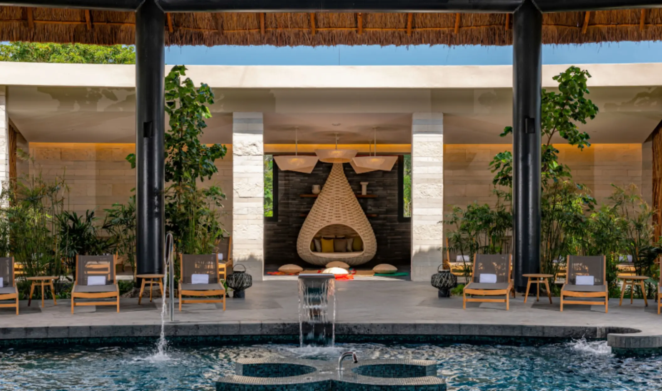 Spa relaxation area with cushioned chairs, lush greenery, and a teardrop-shaped woven daybed by the pool.