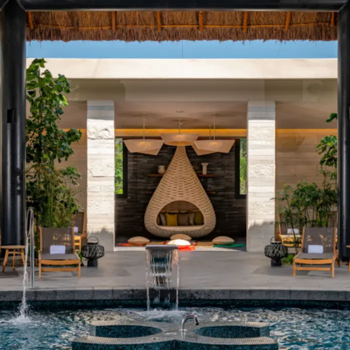 Spa relaxation area with cushioned chairs, lush greenery, and a teardrop-shaped woven daybed by the pool.