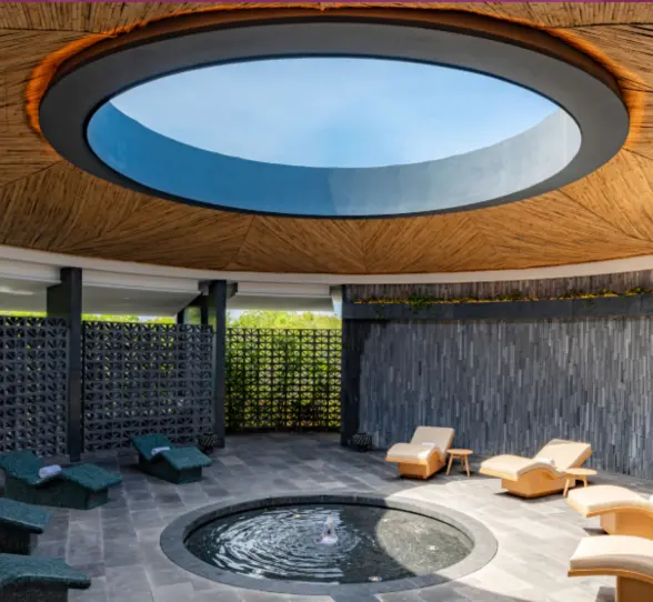 Serene circular spa lounge with water feature, curved loungers, and skylight under a bamboo ceiling.
