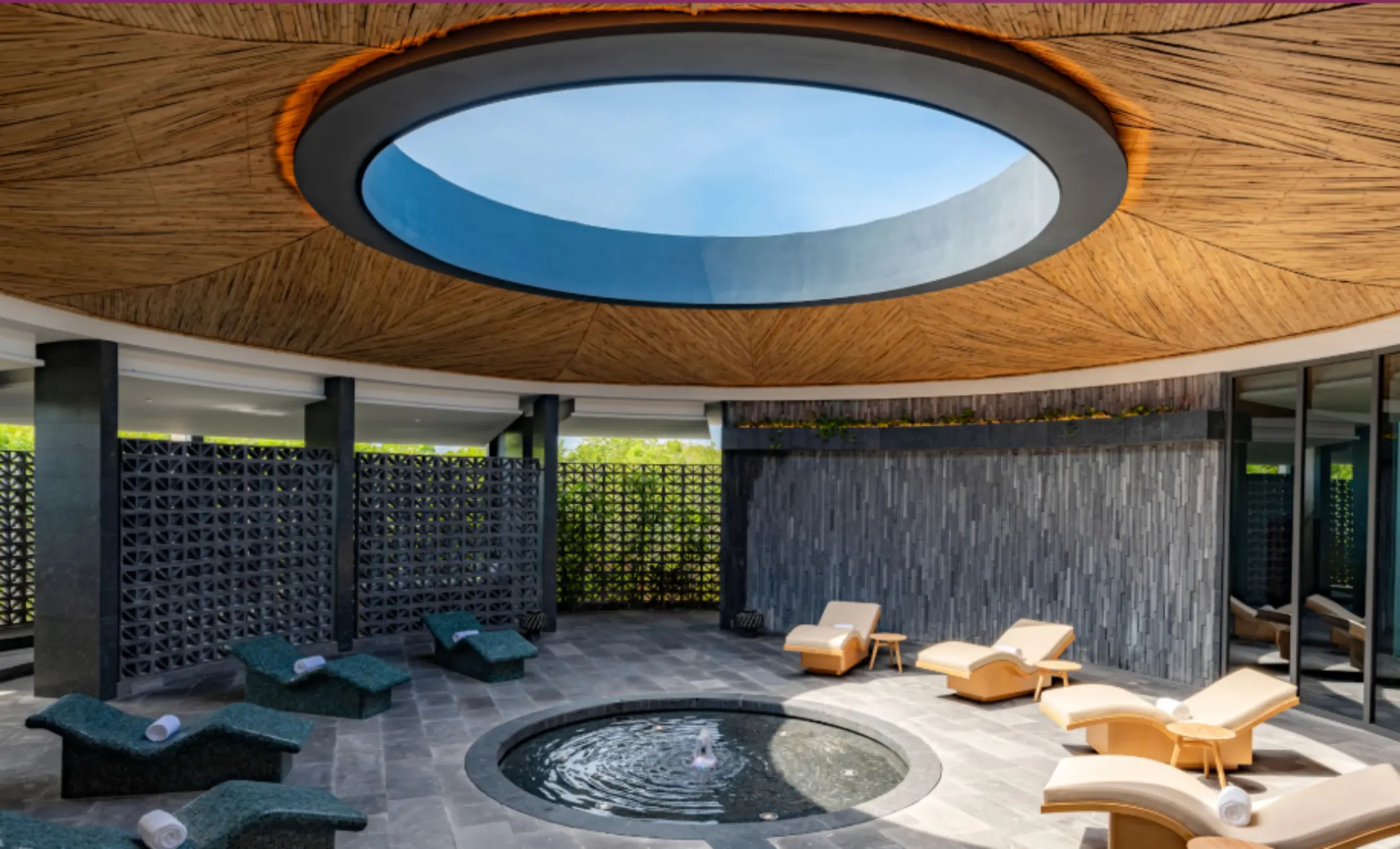 Serene circular spa lounge with water feature, curved loungers, and skylight under a bamboo ceiling.