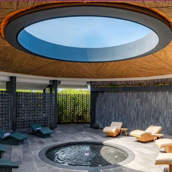 Serene circular spa lounge with water feature, curved loungers, and skylight under a bamboo ceiling.