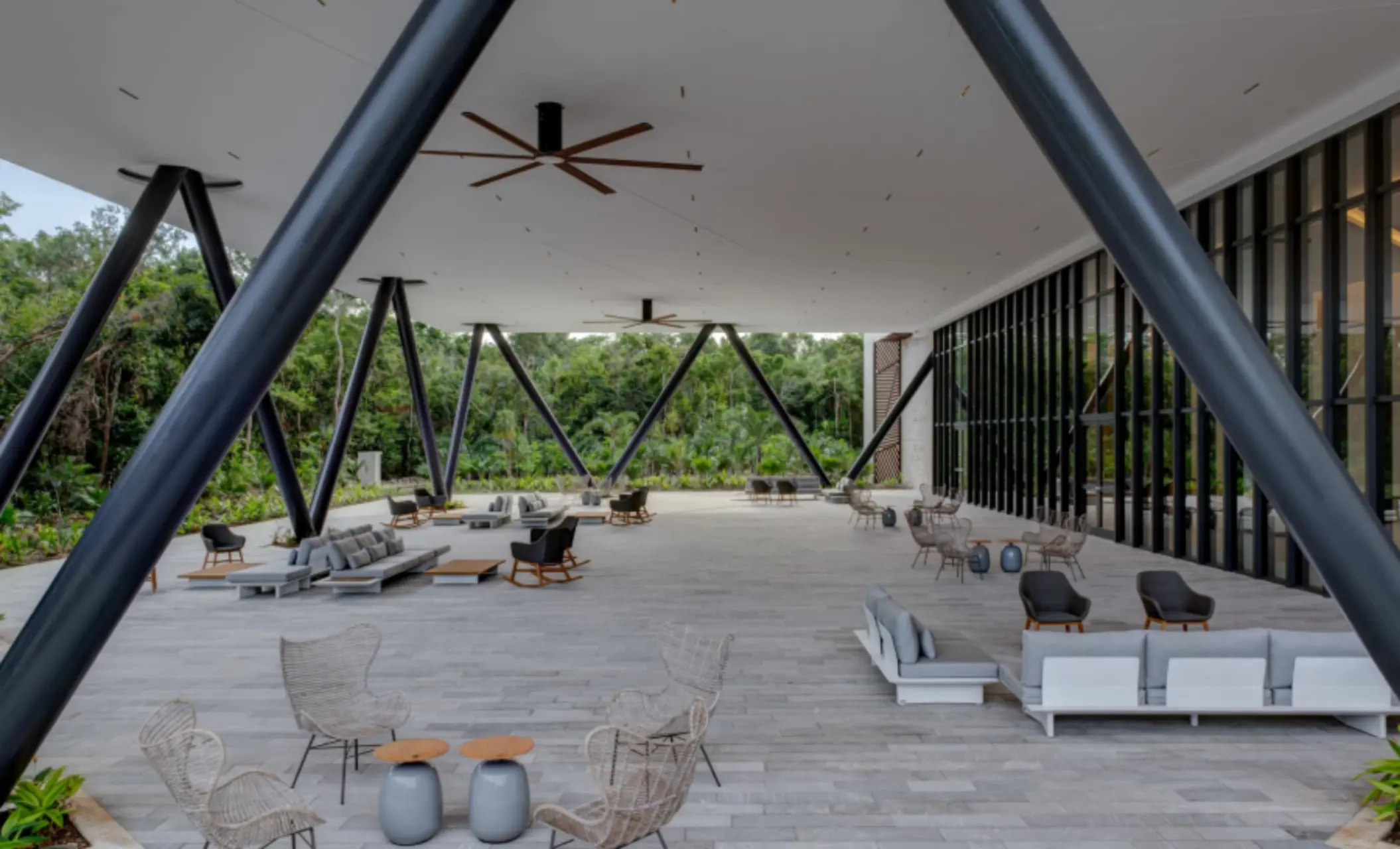 Covered modern terrace lounge surrounded by jungle, featuring neutral sofas, fans, and minimalist decor.
