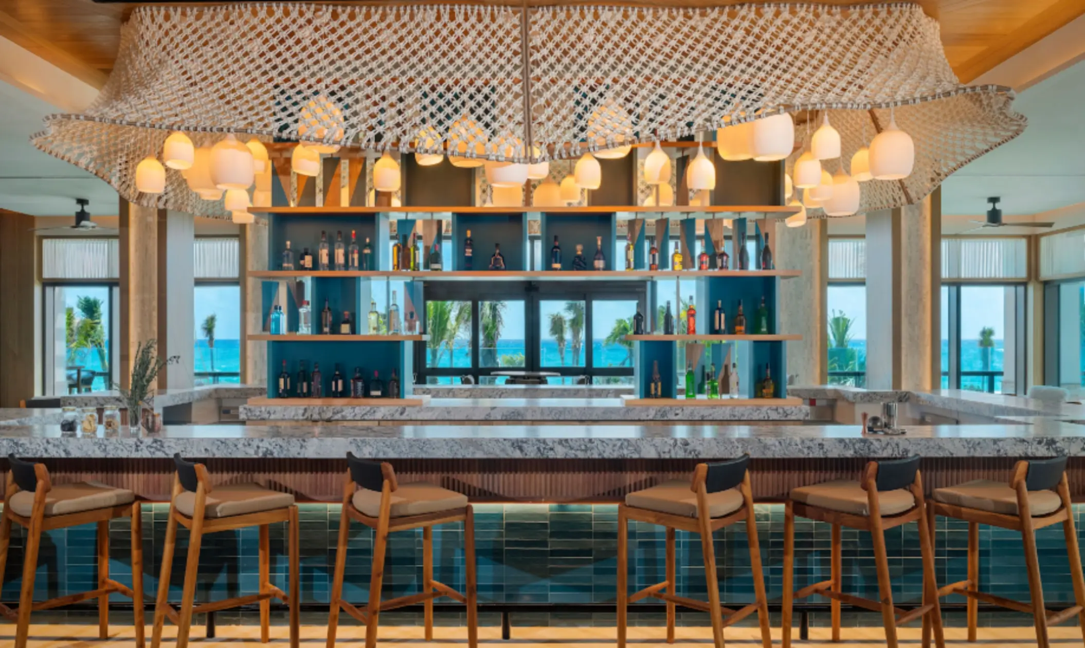 Stylish beachfront bar with ocean views, sleek shelving, marble countertops, and modern hanging lights.