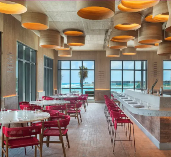 Chic beachfront dining space with ocean views, bold lighting, and red-accented seating in a stylish layout.