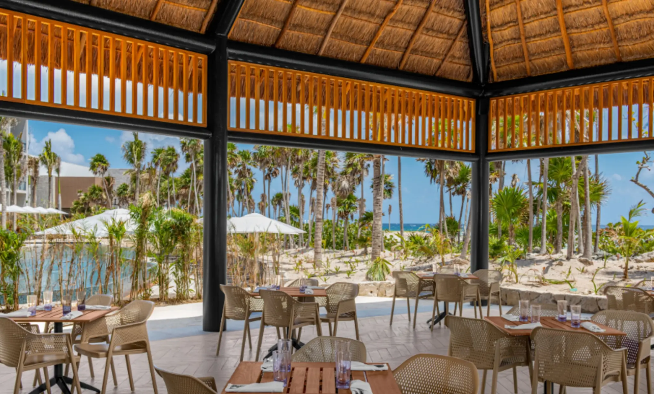 Outdoor thatched-roof dining area with views of palm trees, sandy pathways, and lush tropical greenery.