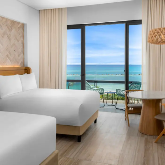 Modern guest room with ocean view balcony, two queen beds, and cozy dining space with wood furniture.
