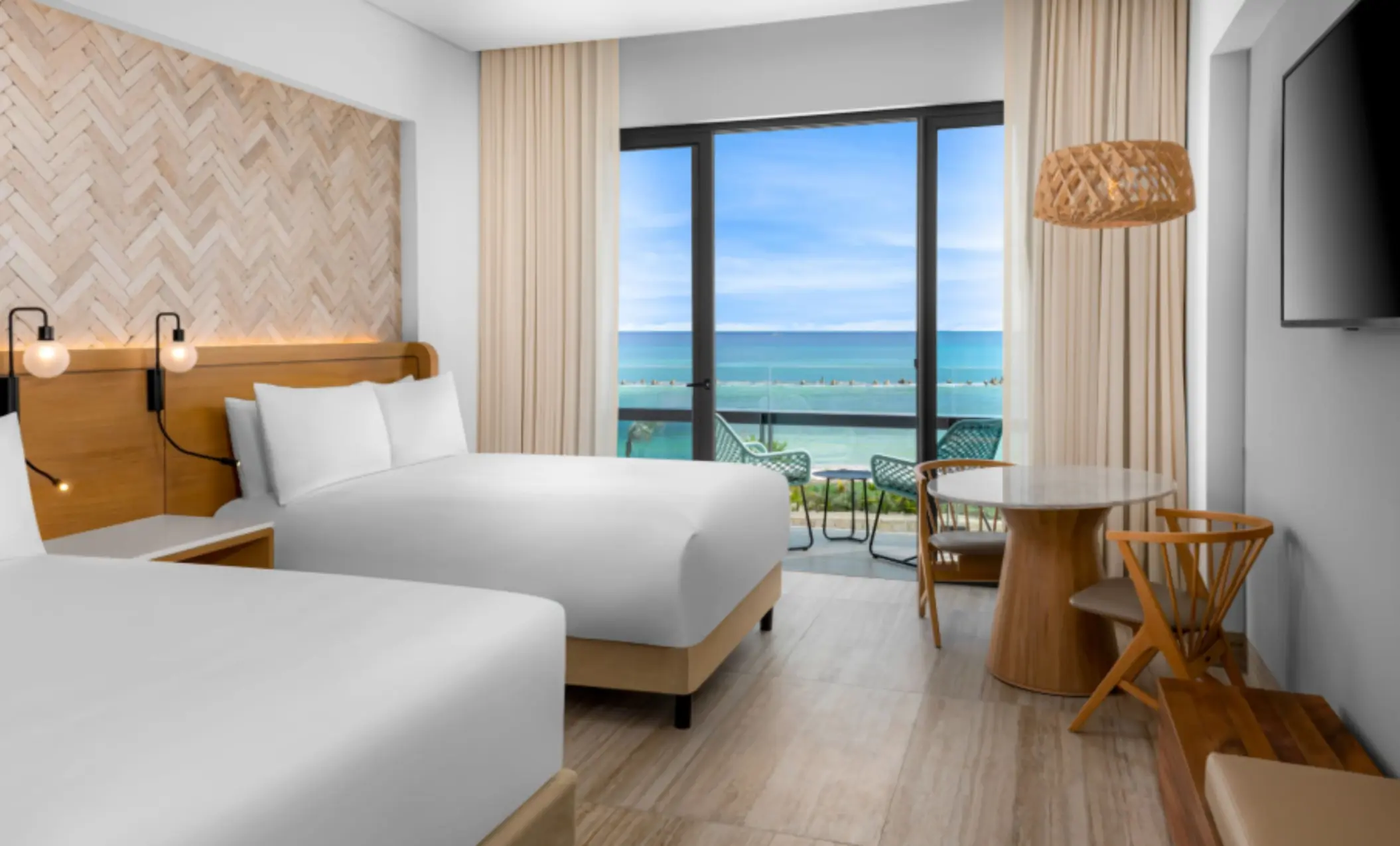 Modern guest room with ocean view balcony, two queen beds, and cozy dining space with wood furniture.