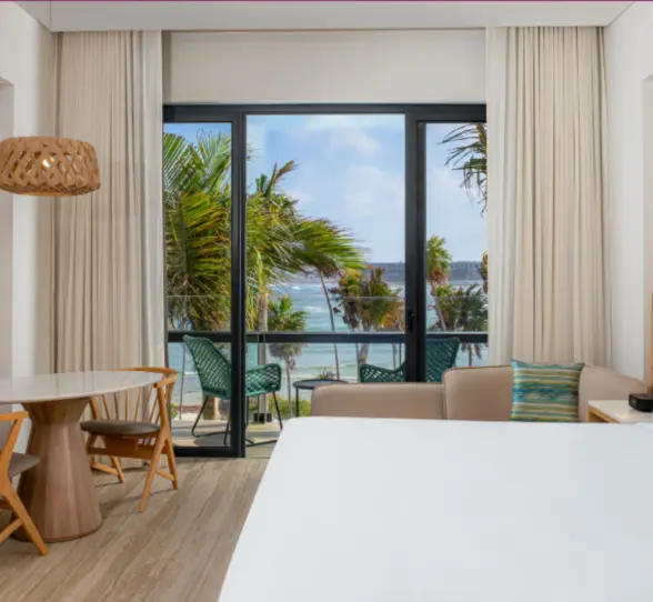 Room with king bed, palm tree and ocean views, natural wood accents, and private balcony seating.