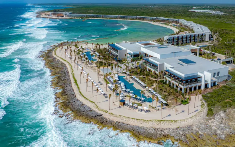 Panoramic aerial shot of beachfront resort with infinity pools, loungers, and dramatic waves below cliffs.