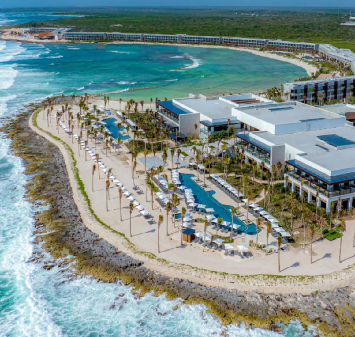 Panoramic aerial shot of beachfront resort with infinity pools, loungers, and dramatic waves below cliffs.
