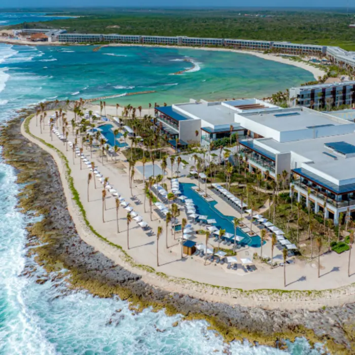 Panoramic aerial shot of beachfront resort with infinity pools, loungers, and dramatic waves below cliffs.