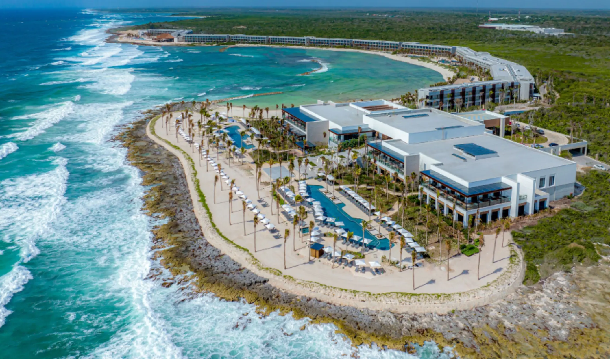 Panoramic aerial shot of beachfront resort with infinity pools, loungers, and dramatic waves below cliffs.