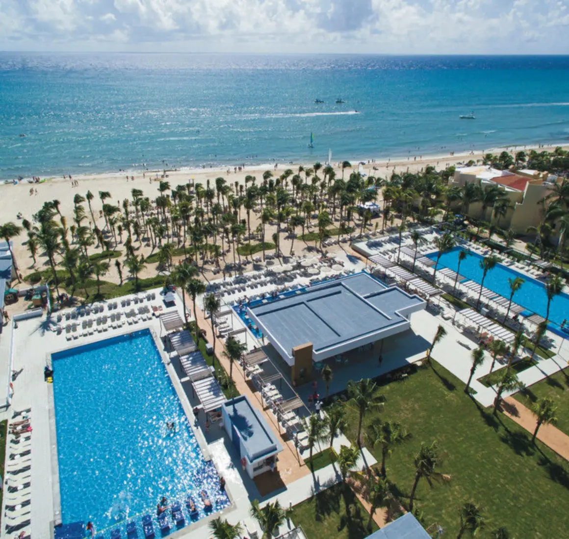 An aerial view showing multiple blue swimming pools, lush palm trees, and a wide sandy beach next to the ocean.