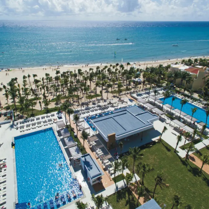 An aerial view showing multiple blue swimming pools, lush palm trees, and a wide sandy beach next to the ocean.