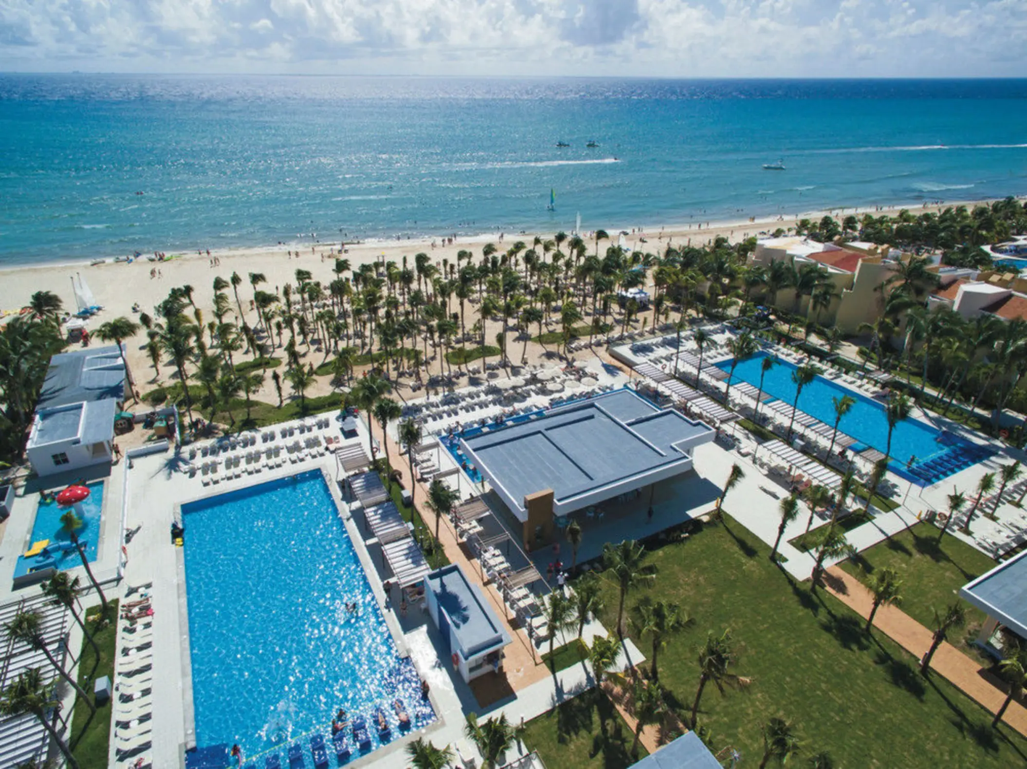 An aerial view showing multiple blue swimming pools, lush palm trees, and a wide sandy beach next to the ocean.