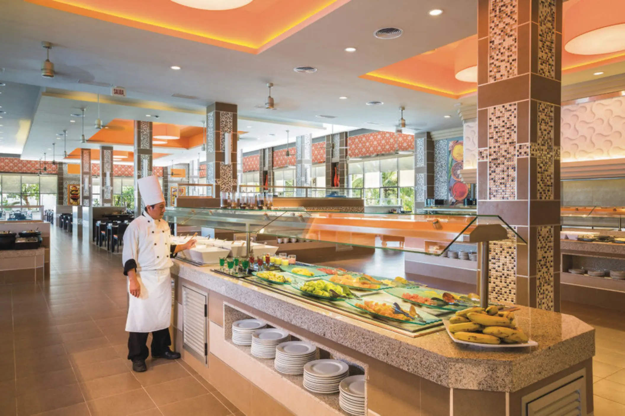 A chef stands next to a vibrant buffet spread with various dishes, in a spacious restaurant with orange ceilings.