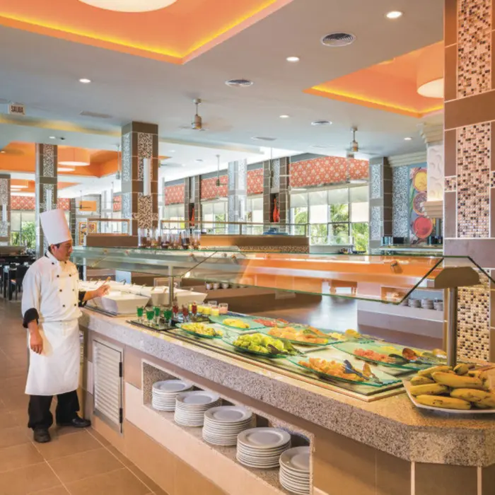 A chef stands next to a vibrant buffet spread with various dishes, in a spacious restaurant with orange ceilings.