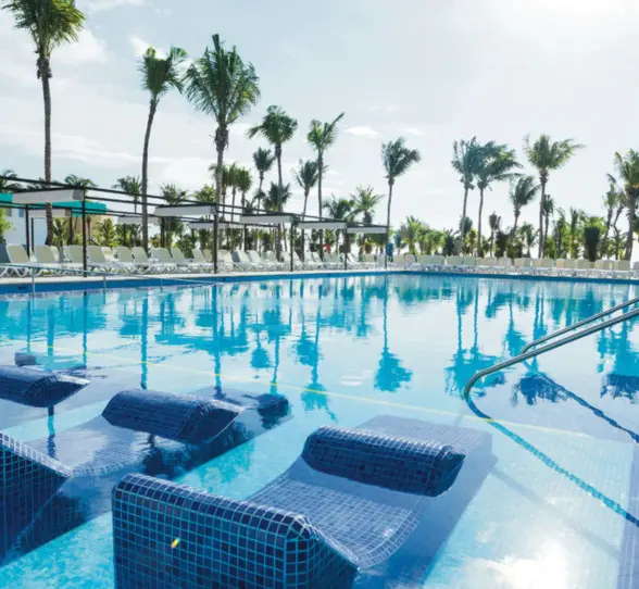 A large outdoor swimming pool with submerged loungers, surrounded by numerous palm trees and sunbeds.