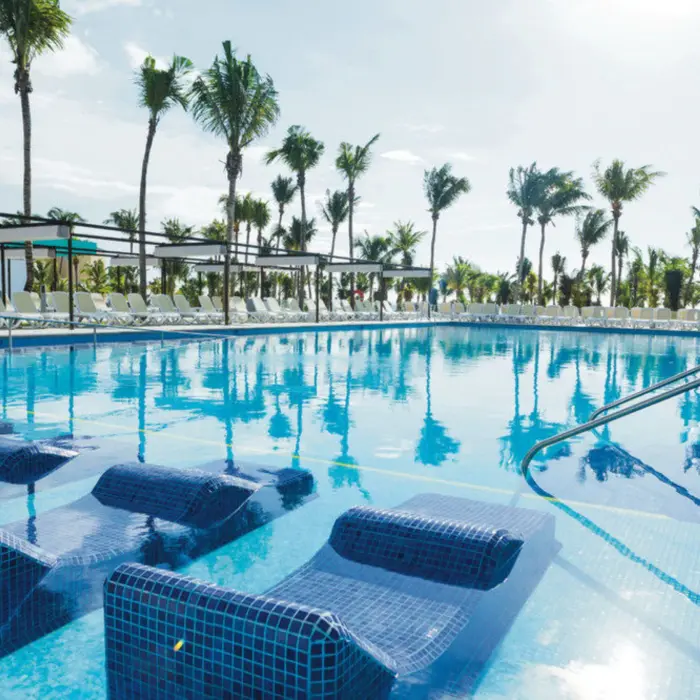A large outdoor swimming pool with submerged loungers, surrounded by numerous palm trees and sunbeds.