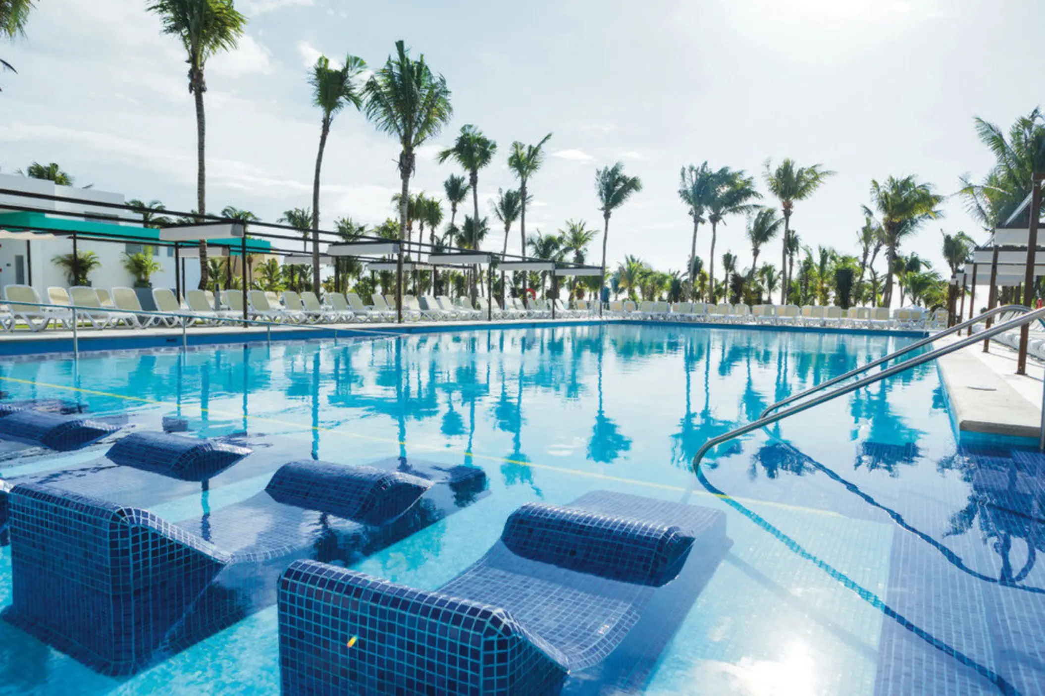 A large outdoor swimming pool with submerged loungers, surrounded by numerous palm trees and sunbeds.