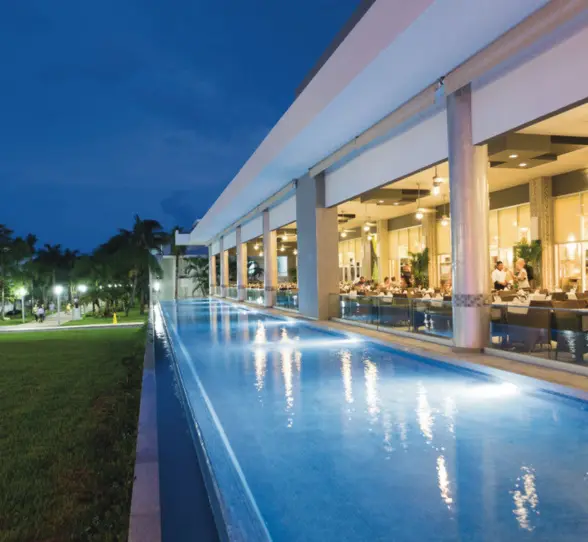 illuminated poolside restaurant at dusk, with diners visible inside and a long, glowing pool alongside a green lawn.