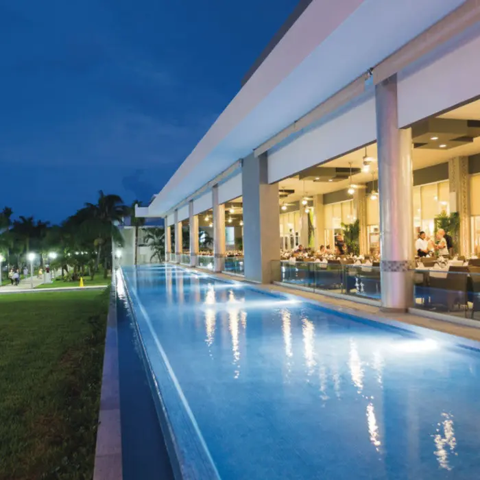 illuminated poolside restaurant at dusk, with diners visible inside and a long, glowing pool alongside a green lawn.