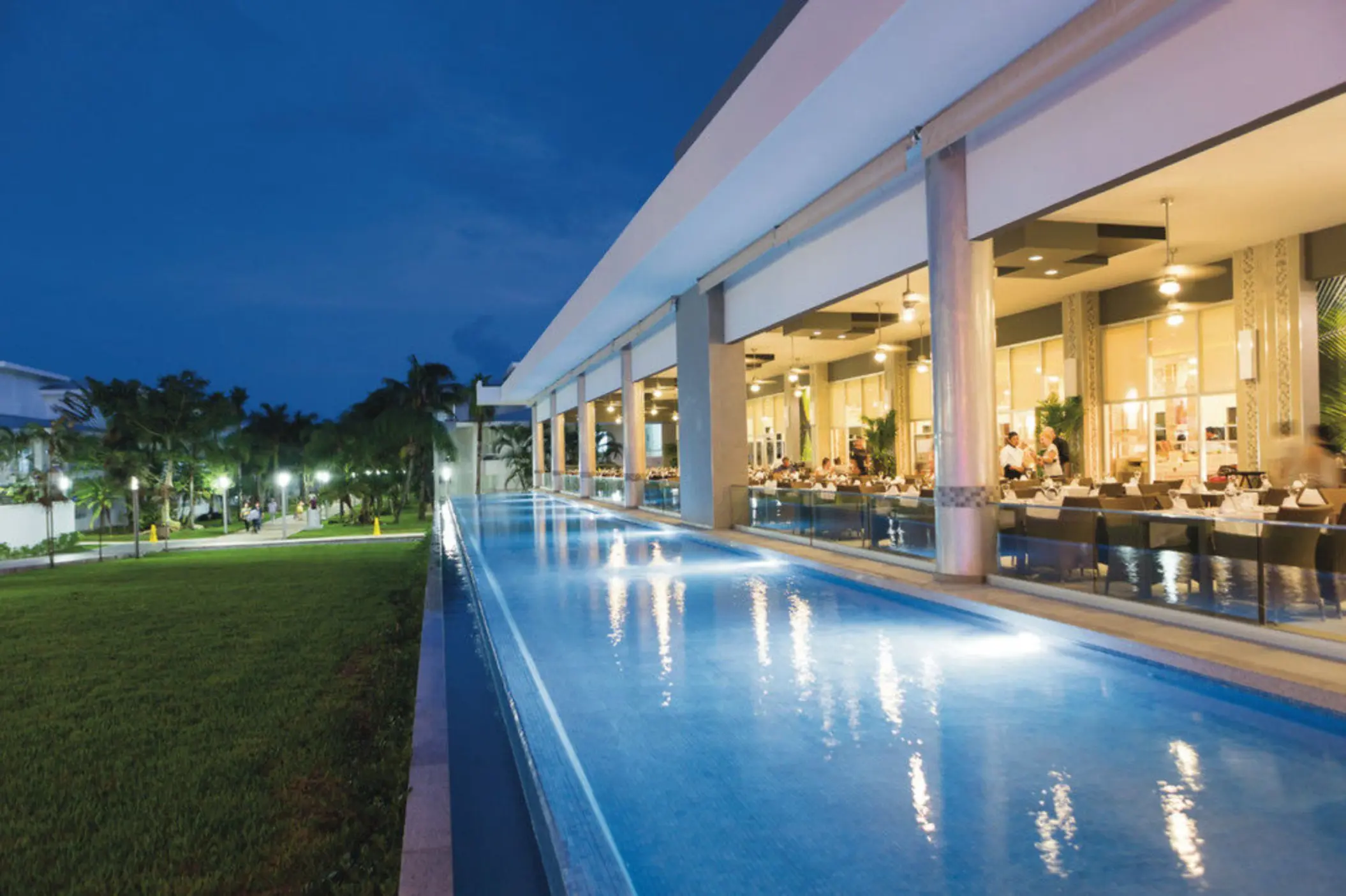 illuminated poolside restaurant at dusk, with diners visible inside and a long, glowing pool alongside a green lawn.