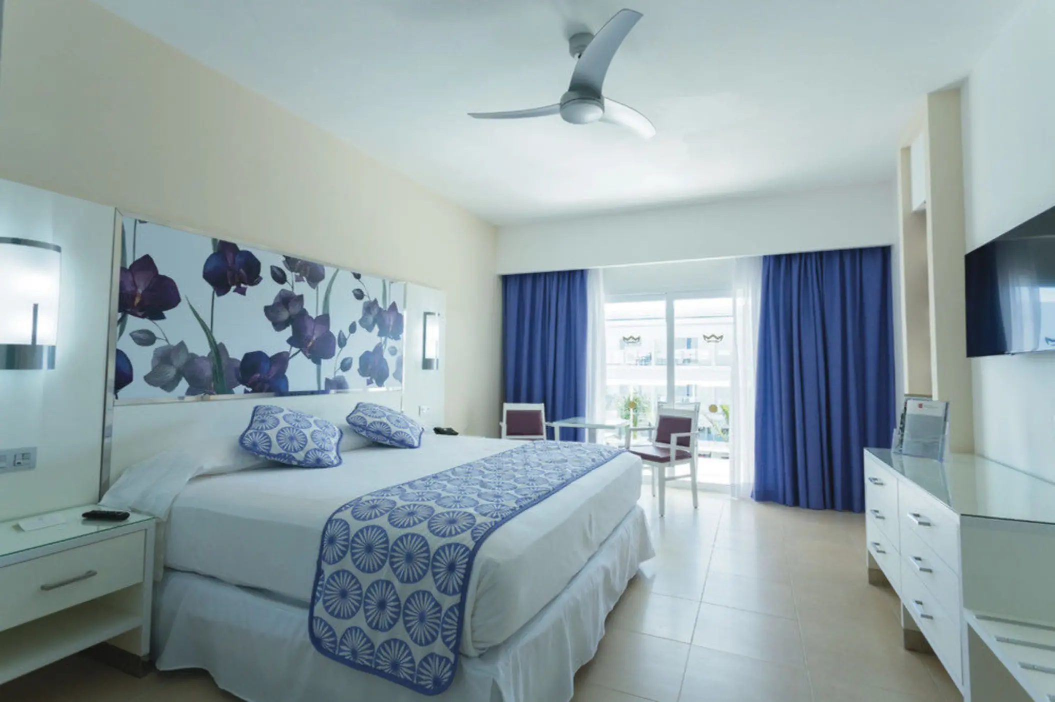 hotel suite with a large bed, purple floral headboard, blue curtains, a ceiling fan, and a flat-screen TV.
