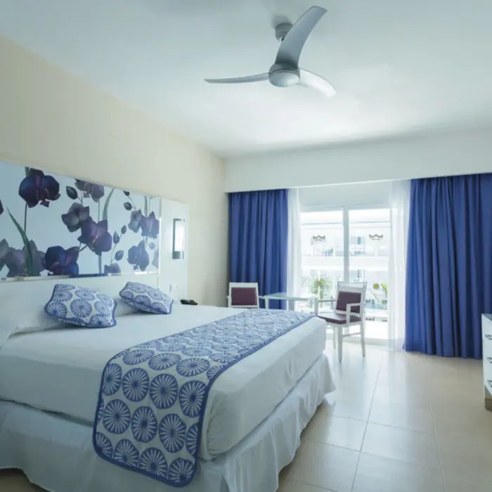 hotel suite with a large bed, purple floral headboard, blue curtains, a ceiling fan, and a flat-screen TV.