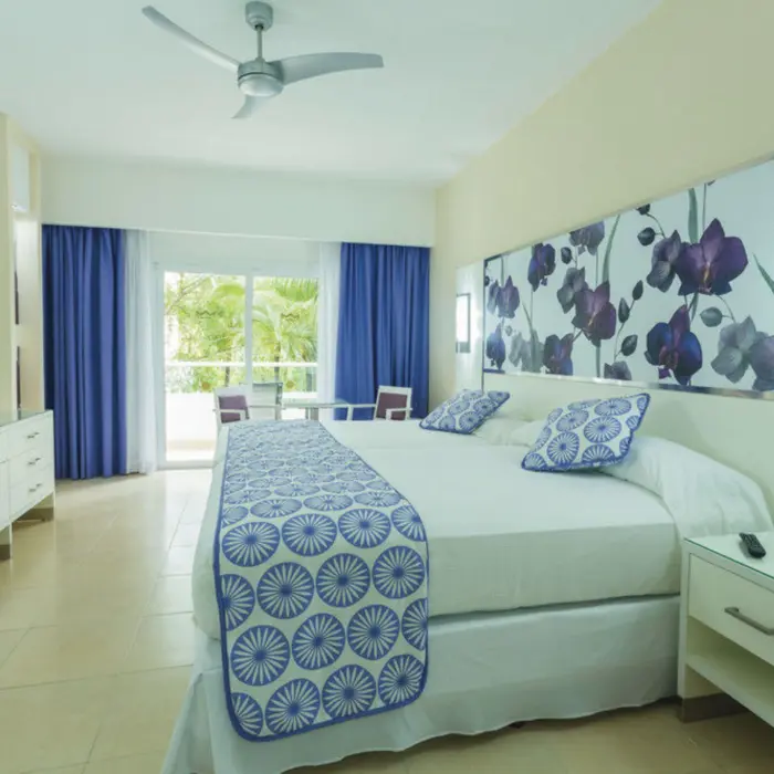 bright suite featuring a large bed with blue and white patterns, a floral headboard, and a balcony with garden views.