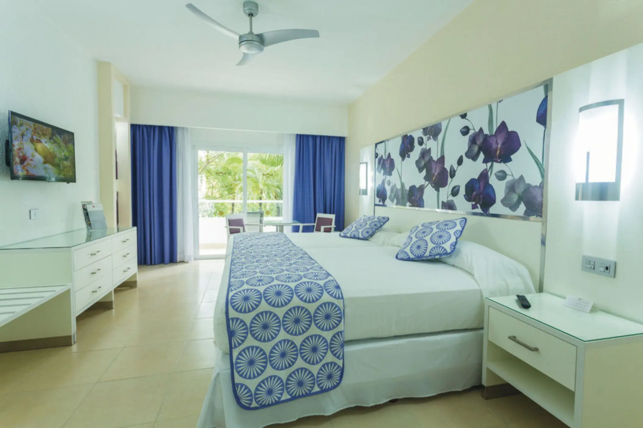 bright suite featuring a large bed with blue and white patterns, a floral headboard, and a balcony with garden views.