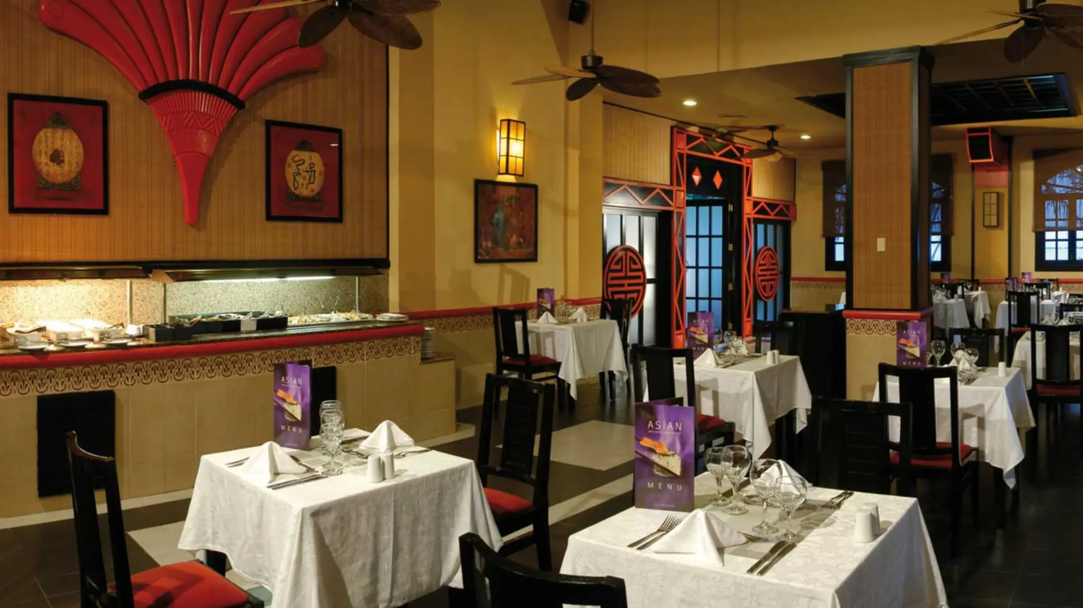 Asian-themed restaurant with red and black decor, traditional art, a buffet counter, and tables set with white tablecloths.