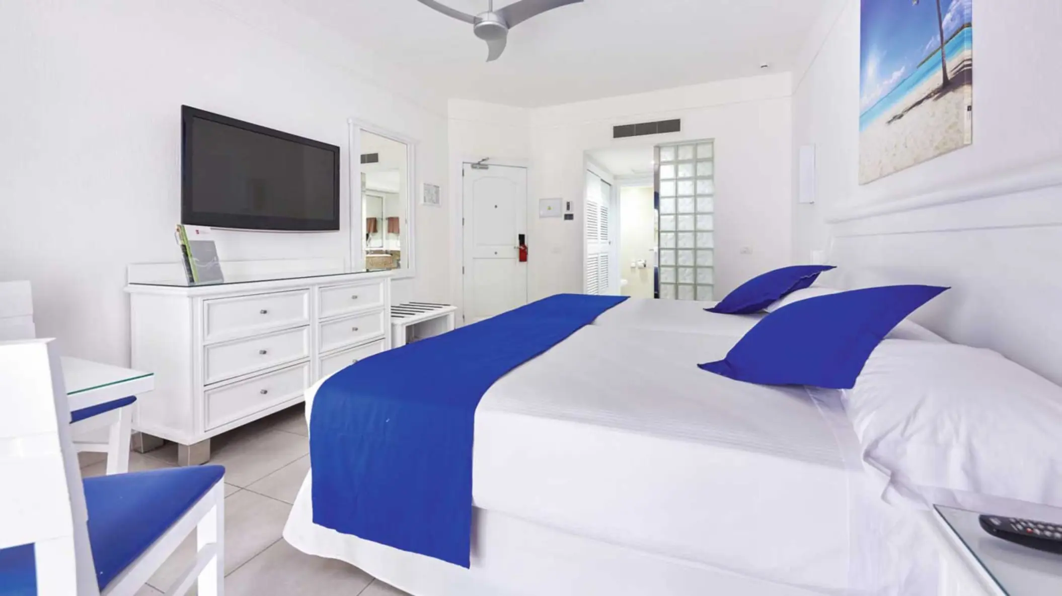 A clean, white connecting double standard room with two beds, blue accents, a TV, and a view into an adjacent room through an open doorway.